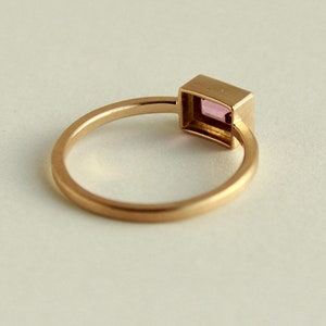 May include: A gold ring with a square-shaped pink gemstone set in a bezel setting.