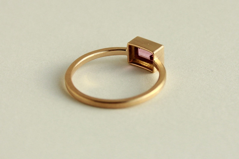 May include: A gold ring with a square-shaped pink gemstone set in a bezel setting.