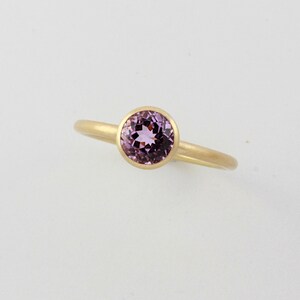 May include: A gold ring with a bezel setting featuring a round, light purple gemstone.