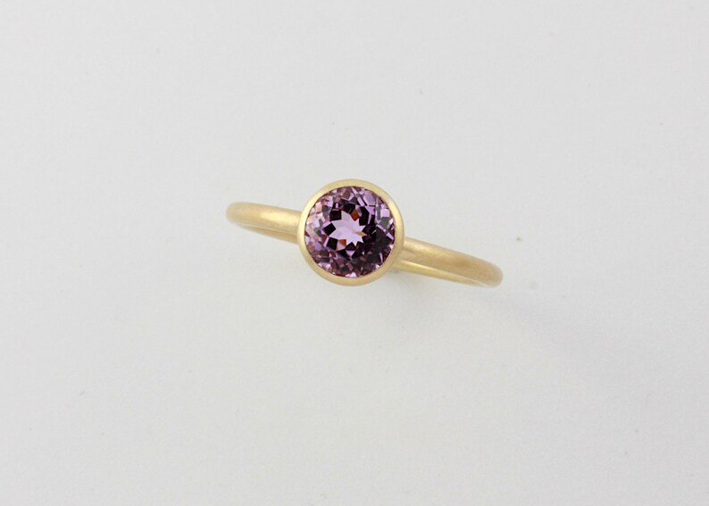 May include: A gold ring with a bezel setting featuring a round, light purple gemstone.