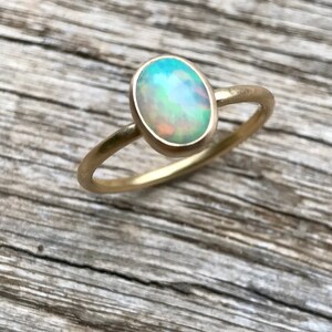 Pu&ograve; includere: Un delicato anello d'oro con una pietra preziosa di opale verde chiaro. L'opale di forma ovale ha una base bianca lattiginosa con riflessi di verde, blu e arancio.