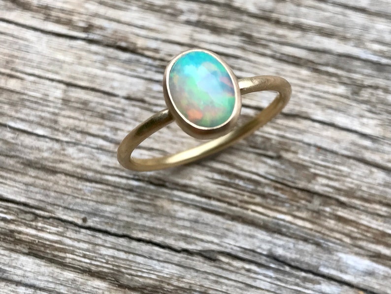 Pu&ograve; includere: Un delicato anello d'oro con una pietra preziosa di opale verde chiaro. L'opale di forma ovale ha una base bianca lattiginosa con riflessi di verde, blu e arancio.