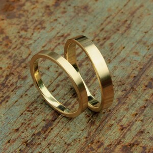 May include: Two gold wedding bands, one slightly larger than the other, are stacked on top of each other on a rustic, brown, textured surface.