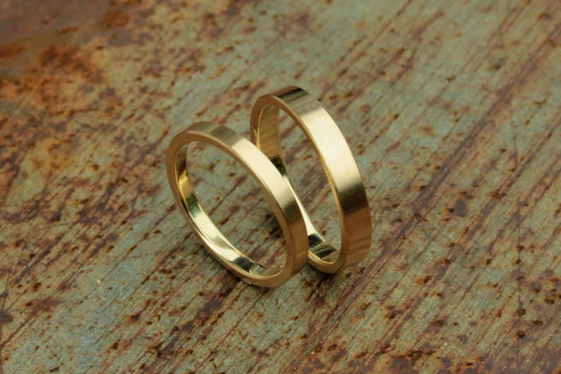 May include: Two gold wedding bands, one slightly larger than the other, are stacked on top of each other on a rustic, brown, textured surface.