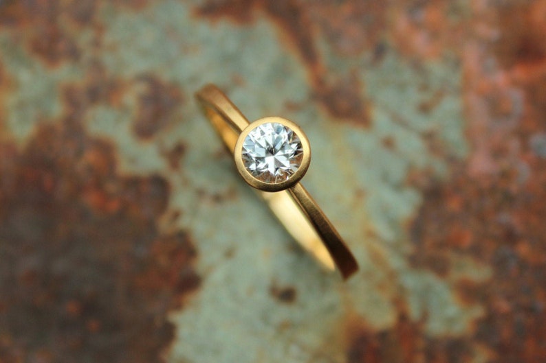 May include: A gold ring with a single round, clear diamond set in a bezel setting.
