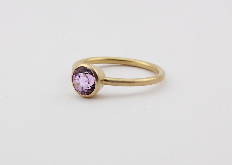 May include: A delicate gold ring with a single round, light purple gemstone set in the center.