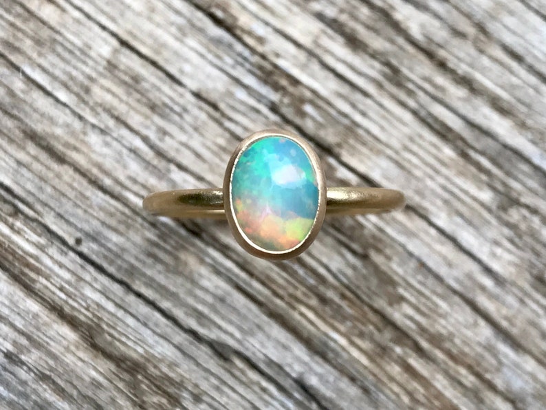 Pu&ograve; includere: Un anello d'oro con una pietra preziosa di opale di forma ovale. L'opale &egrave; di colore blu chiaro con riflessi verdi e arancioni.
