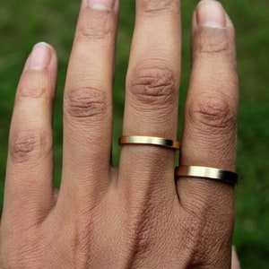 May include: A hand wearing two gold rings. The rings are simple and flat, with a slightly rounded edge.