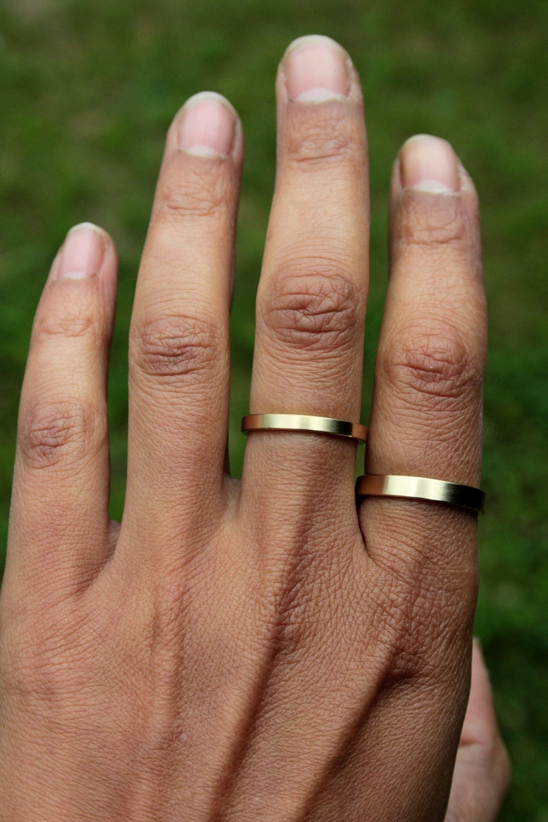 May include: A hand wearing two gold rings. The rings are simple and flat, with a slightly rounded edge.