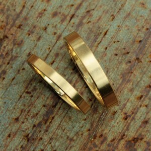 May include: Two gold wedding bands, one slightly wider than the other, are lying on a rustic, brown, and green textured surface.