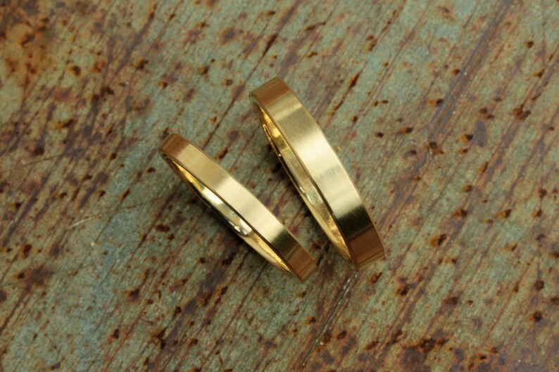 May include: Two gold wedding bands, one slightly wider than the other, are lying on a rustic, brown, and green textured surface.
