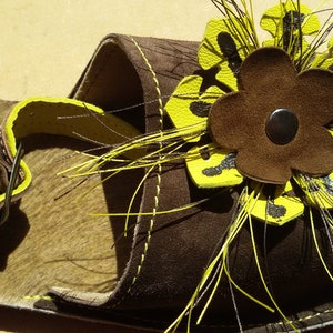 Puede incluir: Sandalias de cuero marr&oacute;n con un adorno de flor amarilla. La flor tiene un centro marr&oacute;n y est&aacute; rodeada de plumas amarillas.