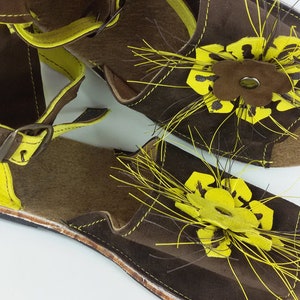 Puede incluir: Un par de sandalias de cuero marr&oacute;n con detalles amarillos y un dise&ntilde;o floral. Las sandalias tienen una correa con hebilla y un detalle de flecos.