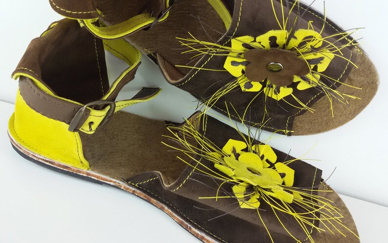 Puede incluir: Un par de sandalias de cuero marr&oacute;n con detalles amarillos y un dise&ntilde;o floral. Las sandalias tienen una correa con hebilla y un detalle de flecos.