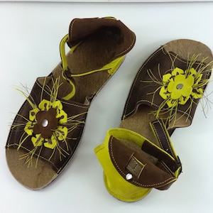 Puede incluir: Un par de sandalias de cuero marr&oacute;n con adornos de flores amarillas. Las sandalias tienen una correa que va alrededor del tobillo y un cierre de hebilla.