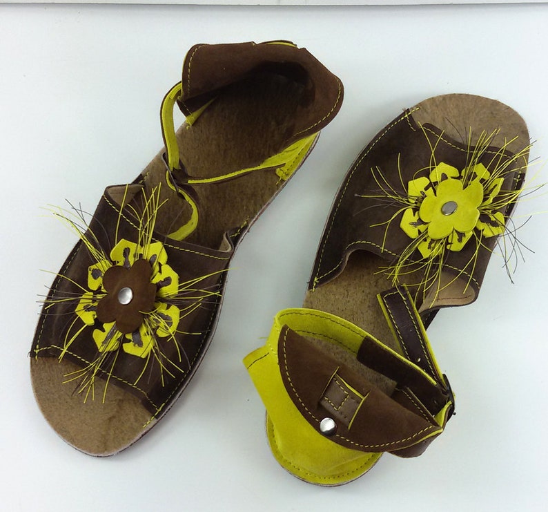 Puede incluir: Un par de sandalias de cuero marr&oacute;n con adornos de flores amarillas. Las sandalias tienen una correa que va alrededor del tobillo y un cierre de hebilla.