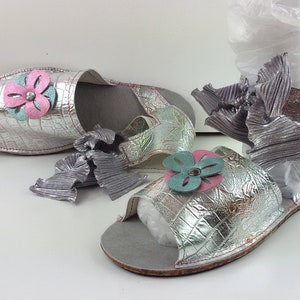 May include: A pair of silver sandals with a flower design. The sandals have a cork sole and a silver strap with a bow.