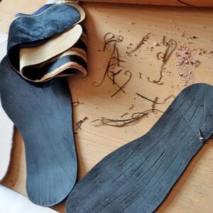Puede incluir: Tres plantillas negras para zapatos, dos son planas y una tiene una superficie texturizada. Las plantillas est&aacute;n sobre una superficie de madera con peque&ntilde;as virutas de madera.