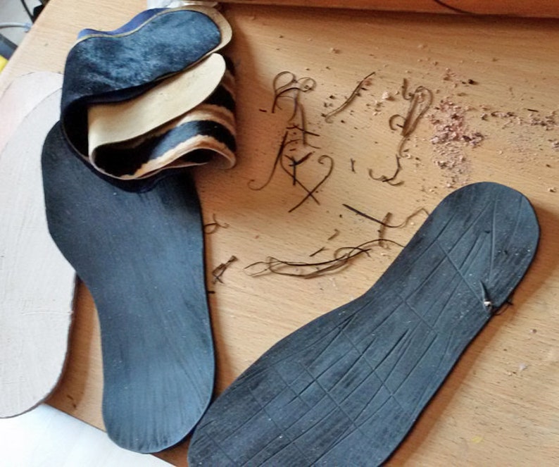 Puede incluir: Tres plantillas negras para zapatos, dos son planas y una tiene una superficie texturizada. Las plantillas est&aacute;n sobre una superficie de madera con peque&ntilde;as virutas de madera.