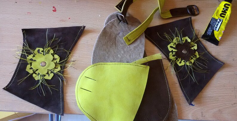 Puede incluir: Primer plano de piezas de cuero marr&oacute;n con detalles en fieltro verde. Las piezas tienen un dise&ntilde;o de flor con un bot&oacute;n marr&oacute;n en el centro. Las piezas est&aacute;n dispuestas sobre una superficie de madera con un tubo de pegamento.