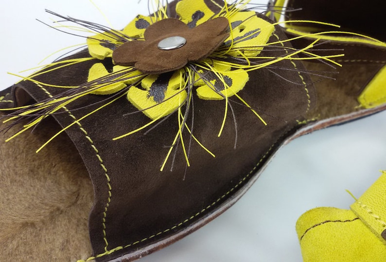 Puede incluir: Sandalias de cuero marr&oacute;n con un adorno de flor amarilla. La flor tiene un centro marr&oacute;n y est&aacute; rodeada de plumas amarillas.