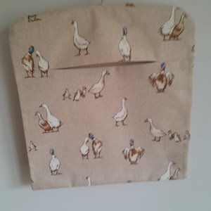 May include: A beige garment bag featuring a repeating pattern of geese in various poses. The geese are depicted in shades of brown, white, and blue. The bag is hanging from a silver metal hanger.