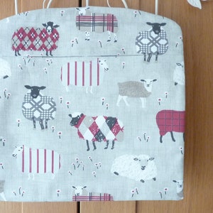 May include: A light gray fabric storage bag featuring a pattern of sheep in various plaid and striped designs. The sheep are in shades of red, gray, and white. The bag has white handles.