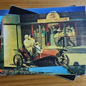 Könnte beinhalten: Eine Vintage-Postkarte mit einem roten und weißen Gebäude mit dem Text "Red Dog Saloon". Das Gebäude hat ein großes Fenster mit einer Gruppe von Menschen im Inneren. Ein Oldtimer steht vor dem Gebäude.