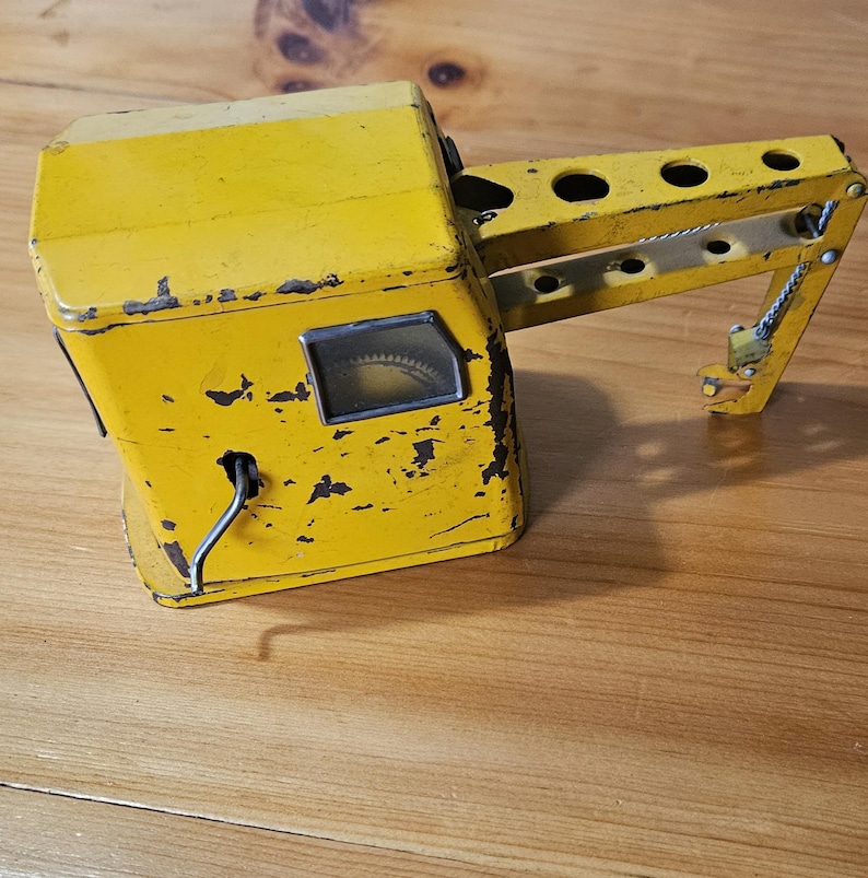 K&ouml;nnte beinhalten: Ein gelber Spielzeugkran aus Metall mit einem Aufziehmechanismus und einem funktionierenden Haken. Der Kran hat ein kleines Fenster mit einem sichtbaren Zahnrad im Inneren. Das Spielzeug weist Gebrauchsspuren auf.