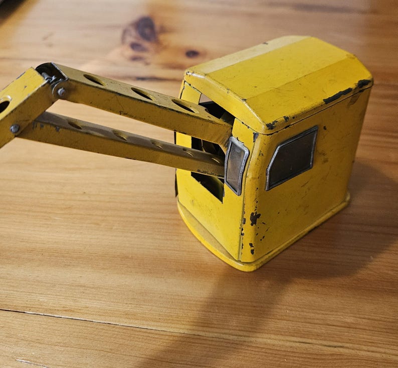 K&ouml;nnte beinhalten: Gelber Spielzeugkran aus Metall mit einem Gelenkarm. Der Kran hat eine kleine Kabine mit Fenstern und ist aus lackiertem Metall.