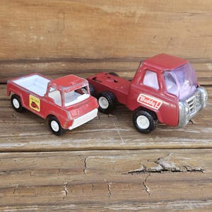 Könnte beinhalten: Zwei rote Vintage-Spielzeug-LKWs. Einer ist ein Pickup mit weißer Ladefläche und einem "Fire Chief"-Aufkleber. Der andere ist ein Sattelzug mit "Buddy L"-Logo. Beide haben schwarze Reifen und weiße Felgen.