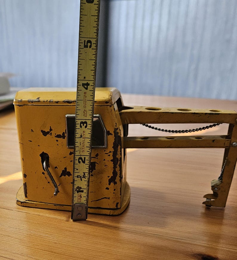 K&ouml;nnte beinhalten: Ein gelber Spielzeugkran mit einer Metallkette und einem Hebel. Der Kran ist etwa 13 cm hoch und aus Metall. Er hat einen abgenutzten, Vintage-Look.