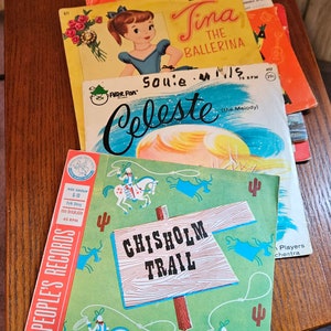 May include: A stack of vintage children's records with colorful covers. The top record features a cowboy riding a horse on a green background with the text "Chisholm Trail". The record below features a ballerina with the text "Tina The Ballerina". The bottom record features a girl with the text "Souie Mills Celeste".