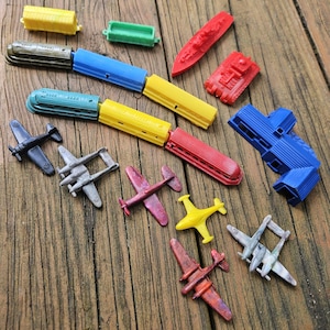Könnte beinhalten: Eine Sammlung von Miniatur-Spielzeugen aus Kunststoff, darunter zwei mehrfarbige Spielzeugeisenbahnen, mehrere kleine Flugzeuge in verschiedenen Farben, ein roter Spielzeugpanzer, ein rotes Schiff, ein blaues Gebäude und zwei kleine rechteckige Waggons.