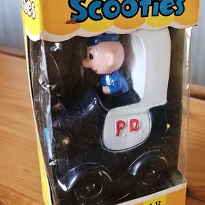 Könnte beinhalten: Ein schwarzes und weißes Aufziehspielzeug-Polizeiauto mit einem Cartoon-Polizisten am Steuer. Das Auto hat ein 'PD'-Logo an der Seite und ist in einer gelben Schachtel verpackt, auf der 'Cragstan Scooties' und 'Walking Jalopies' steht.