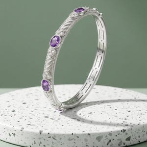 Könnte beinhalten: Ein silberfarbener Klapparmreif mit einem texturierten, gedrehten Metallband und drei oval geschliffenen violetten Amethysten. Das Armband ist mit kleinen, klaren, runden Steinen verziert, die in blumenförmigen Metallfassungen sitzen.