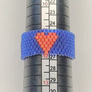 May include: A blue beaded ring with a red heart design. The ring is being measured on a sizing tool, showing a size of 27.