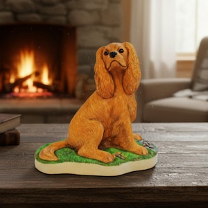 Op de afbeelding: Een goudbruin spaniel-beeldje in een zittende houding. De hond heeft lange, getextureerde oren, een donkere neus en expressieve ogen. Het staat op een gebeeldhouwde basis die is beschilderd als een stukje groen gras met bruine details.