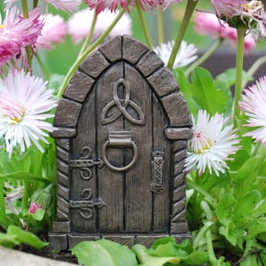 Peut inclure: Une porte de fée cintrée marron avec une finition en grain de bois texturé, un heurtoir circulaire central, deux charnières métalliques décoratives à gauche, une poignée verticale à droite et un symbole de nœud triquetra sculpté au-dessus.