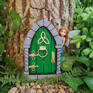 Peut inclure: Une porte miniature arquée verte avec un heurtoir en anneau doré, des charnières dorées et un emblème de nœud celtique doré. Une petite figurine de fée aux cheveux orange et aux ailes translucides se tient à droite du cadre en pierre grise.