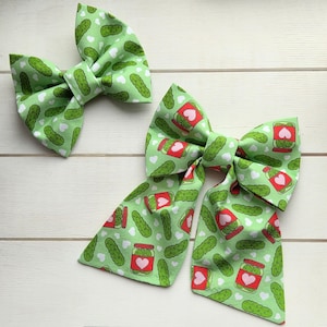 May include: Two green fabric hair bows. One bow features a pattern of pickles and white hearts. The other bow has a pattern of pickles, white hearts, and jars with hearts. The bows are on a white wooden surface.