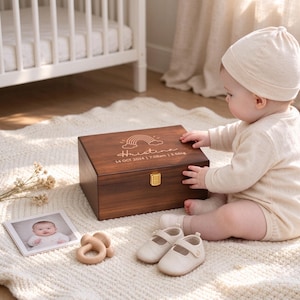 May include: A rectangular dark wood keepsake box with a gold latch, engraved with a rainbow, the name Hristina, and the birth details 14 OCT 2024 | 7:08am | 3.56kg. The box is placed on a textured white blanket near baby shoes and a photo.