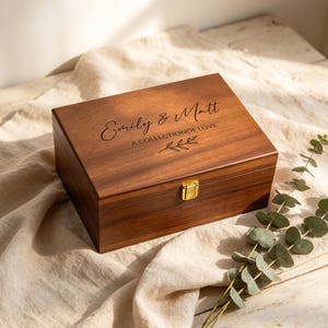 May include: A rectangular wooden keepsake box with a dark brown finish and a gold-colored metal latch. The lid is engraved with the text Emily & Matt, A COLLECTION OF LOVE, and a small leaf graphic. The box has a smooth, polished wood grain surface.
