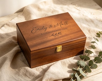 May include: A rectangular wooden keepsake box with a dark brown finish and a gold-toned metal latch. The lid is engraved with the text Emily & Matt, A COLLECTION OF LOVE, and a small leaf sprig design. The box has a smooth, polished surface.