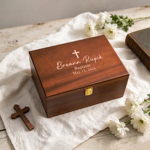 May include: A rectangular dark wood keepsake box with a gold-tone metal latch. The lid is engraved with a cross symbol and the text Breann Rupik, Baptism, May 13, 2026. A small wooden cross and white flowers are placed nearby on a white fabric.