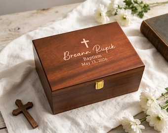 May include: A rectangular dark wood keepsake box with a gold-tone metal latch. The lid is engraved with a cross symbol and the text Breann Rupik, Baptism, May 13, 2026. A small wooden cross and white flowers are placed nearby on a white fabric.