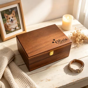 May include: A rectangular wooden keepsake box with a gold-colored metal latch. The lid is engraved with the name Olivia, the dates 2008-2023, and two small paw prints. The box features a smooth, dark brown wood grain finish and a hinged lid.