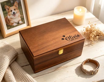 May include: A rectangular wooden keepsake box with a gold-coloured metal latch. The lid is engraved with the name Olivia, the dates 2008-2023, and two small paw prints. The box features a smooth, dark brown wood grain finish and a hinged lid.