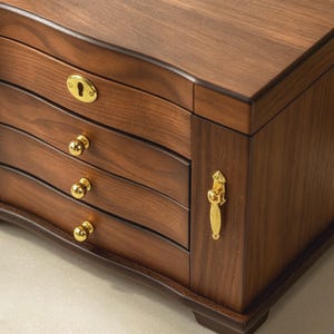 May include: A dark brown wooden jewellery box featuring three horizontal drawers with small round gold knobs, a gold keyhole plate on the top drawer, and a vertical gold handle on the side panel, showcasing a smooth wood grain finish.