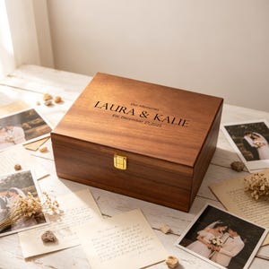 Puede incluir: Una caja de recuerdos rectangular de madera con tapa abatible y un cierre de metal dorado. La tapa está grabada con el texto "Our Memories", "LAURA & KALIE" y "Est. December 27, 2025". La caja presenta un acabado de madera de color marrón oscuro.